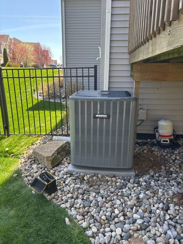 Completed full air conditioning system installation at residential property. New AC condenser unit installed on existing concrete pad with river rock landscaping, positioned adjacent to home exterior near deck structure. System properly mounted, refrigerant lines connected and pressure-tested per manufacturer specifications, electrical connections completed with appropriate safety labeling in place, and condensate drainage system installed and operational.