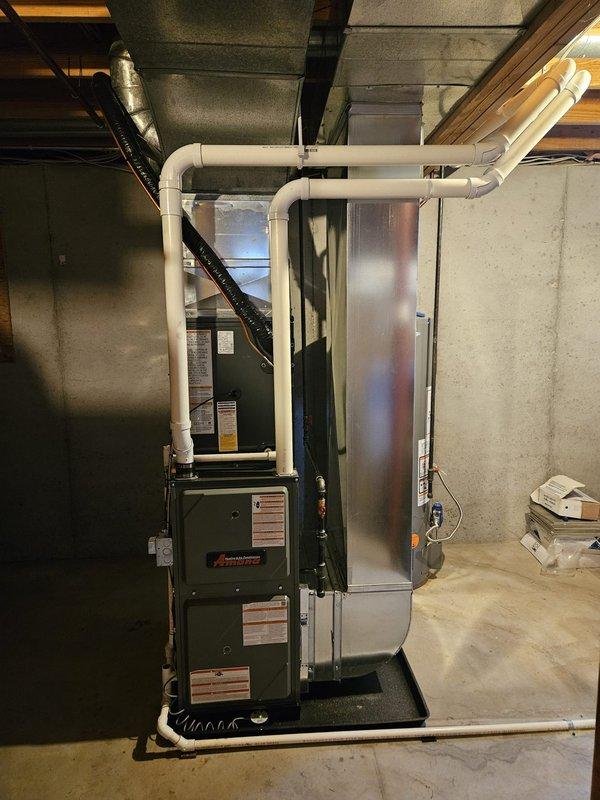 Complete HVAC system replacement performed, including installation of new furnace and air conditioning unit. Furnace installed in basement utility area with air handler positioned adjacent, connected via white PVC condensate drainage system and integrated black flexible ductwork. Outdoor condenser unit installed on stone base against exterior wall with proper refrigerant line connections, electrical disconnect box, and service access. System charged, tested, and verified operational upon completion.