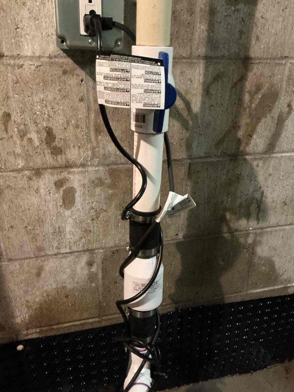 Serviced existing sump pump system in basement utility area. Inspected vertical PVC discharge pipe assembly secured to concrete block wall with proper mounting straps, verified electrical connection at wall-mounted outlet box, and confirmed operational status of pump within concrete pit. System found functioning properly with discharge piping, check valve, and drainage components in good working condition.