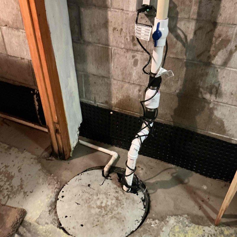 Serviced existing sump pump system in basement utility area. Inspected vertical PVC discharge pipe assembly secured to concrete block wall with proper mounting straps, verified electrical connection at wall-mounted outlet box, and confirmed operational status of pump within concrete pit. System found functioning properly with discharge piping, check valve, and drainage components in good working condition.