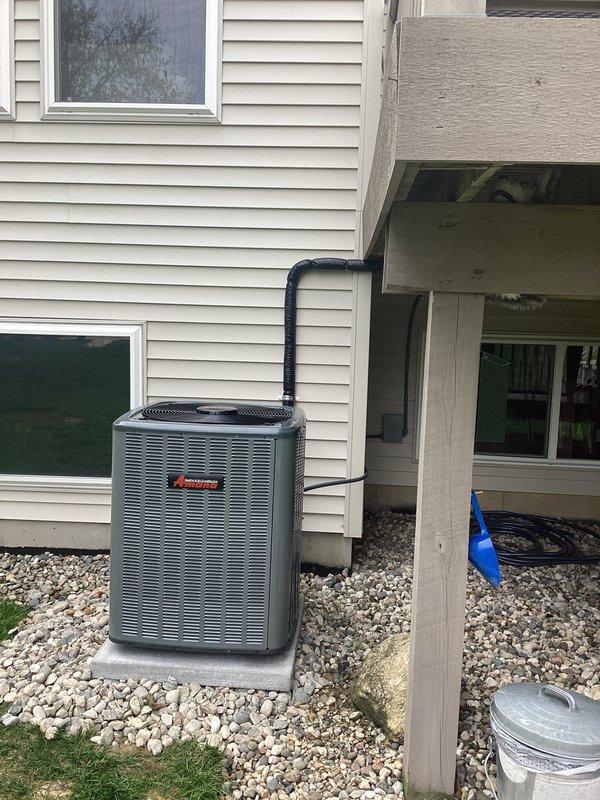 Completed full residential air conditioning system installation at single-family home with cream-colored vinyl siding. Installed new Amana brand outdoor condensing unit on existing concrete pad with proper gravel landscaping base for drainage and equipment support. Routed black insulated refrigerant line set from condenser unit vertically along exterior wall siding to connect with indoor air handler. Positioned equipment near foundation with adequate clearance for airflow and future service access. Unit installed under elevated deck structure with sufficient overhead clearance for proper ventilation and maintenance accessibility. All refrigerant lines properly insulated and secured to building exterior. System installation completed with equipment operational and ready for use.