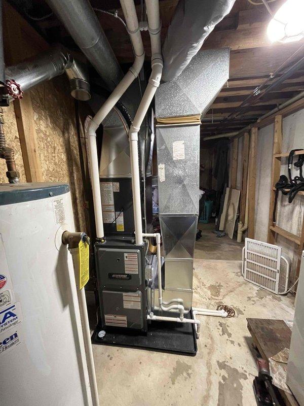 Completed full residential HVAC system installation including new furnace and air conditioning unit. Furnace installed in unfinished basement utility area with proper venting via white PVC piping, connected to existing ductwork system, and mounted on code-compliant base platform. Exterior AC condenser unit positioned on building exterior near electrical meter with appropriate clearances maintained. All refrigerant lines, electrical connections, and condensate drainage properly installed and tested for system functionality.