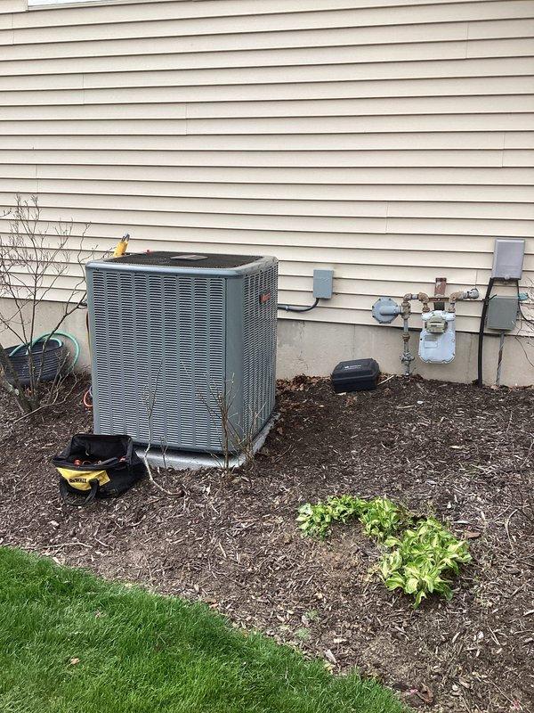 Completed full air conditioning system replacement at residential property. Removed existing outdoor condenser unit and installed new gray metal AC condenser unit on exterior wall with beige vinyl siding, positioned on mulched landscaping area. Ran new refrigerant lines (copper and insulated) from condenser to building with proper mounting brackets and hangers secured to exterior wall. Connected all electrical and refrigerant connections, performed vacuum and pressure testing of system, and charged unit to manufacturer specifications. Verified all utility connections including proximity to gas meter assembly were properly maintained. System tested and operational upon completion.