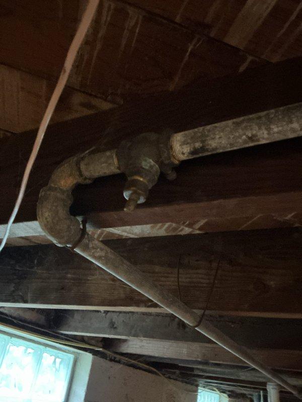 Conducted project estimate for replacement of failed water shutoff valve in 75-year-old residential property. The existing valve is broken and stuck in the open position, preventing proper water control and blocking a city-mandated water meter replacement. Documented heavily corroded supply piping in basement with significant deterioration evident on metal pipes throughout the system. Photographed current plumbing assembly including existing water meter with brass fittings showing oxidation and patina, along with deteriorated insulation on pipe connections. Assessed structural conditions of basement area with exposed floor joists and evaluated accessibility for valve replacement work required to facilitate upcoming municipal water meter upgrade.