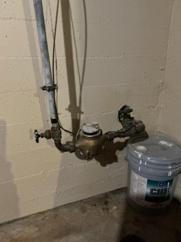 Conducted project estimate for replacement of failed water shutoff valve in 75-year-old residential property. The existing valve is broken and stuck in the open position, preventing proper water control and blocking a city-mandated water meter replacement. Documented heavily corroded supply piping in basement with significant deterioration evident on metal pipes throughout the system. Photographed current plumbing assembly including existing water meter with brass fittings showing oxidation and patina, along with deteriorated insulation on pipe connections. Assessed structural conditions of basement area with exposed floor joists and evaluated accessibility for valve replacement work required to facilitate upcoming municipal water meter upgrade.
