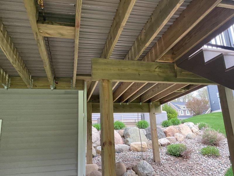 Conducted site assessment for outdoor natural gas line servicing gas grill that requires modification due to new deck construction on 6-year-old residential property. Evaluated existing gas line routing beneath newly installed elevated deck structure with pressure-treated lumber framing and corrugated metal ceiling panels. Documented current gas line configuration and determined necessary repairs and/or extension requirements to accommodate deck installation while maintaining proper clearances and code compliance. Photographed underside of deck construction showing wooden beam framework, support posts, and landscaped area with decorative rock groundcover to establish scope of work for gas line relocation or extension project.