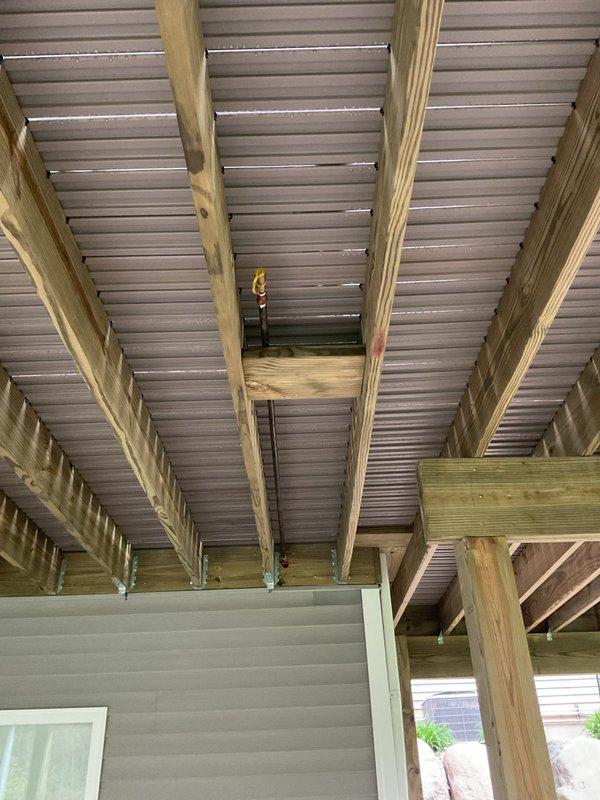 Conducted site assessment for outdoor natural gas line servicing gas grill that requires modification due to new deck construction on 6-year-old residential property. Evaluated existing gas line routing beneath newly installed elevated deck structure with pressure-treated lumber framing and corrugated metal ceiling panels. Documented current gas line configuration and determined necessary repairs and/or extension requirements to accommodate deck installation while maintaining proper clearances and code compliance. Photographed underside of deck construction showing wooden beam framework, support posts, and landscaped area with decorative rock groundcover to establish scope of work for gas line relocation or extension project.