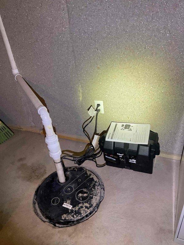 Replaced battery backup sump pump system in basement utility area. Installed new sump pump with black circular basin cover, white PVC discharge piping with multiple joints and fittings routed along wall, and black electrical control box with proper wiring to wall outlet. System tested and operational upon completion.