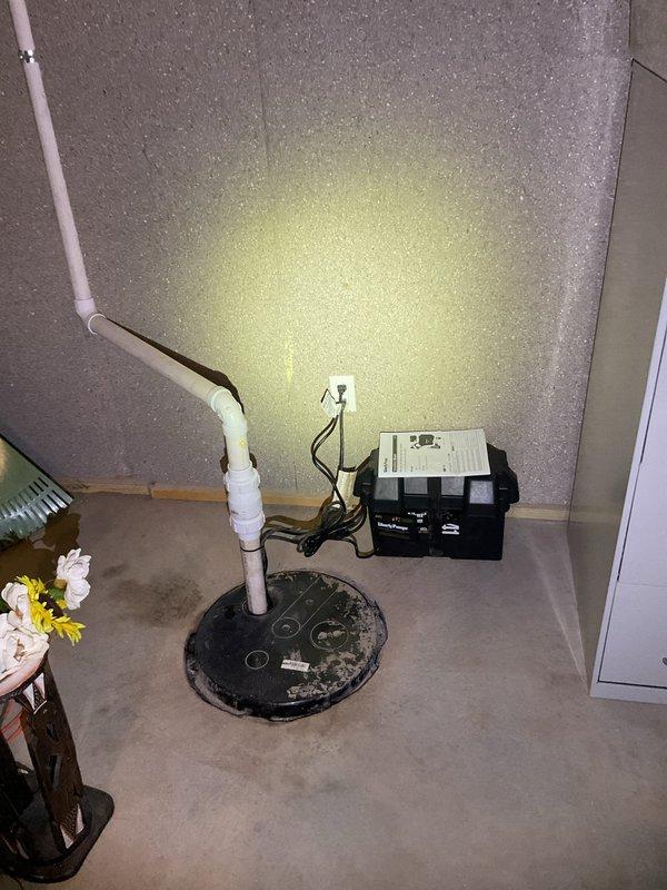 Replaced battery backup sump pump system in basement utility area. Installed new sump pump with black circular basin cover, white PVC discharge piping with multiple joints and fittings routed along wall, and black electrical control box with proper wiring to wall outlet. System tested and operational upon completion.