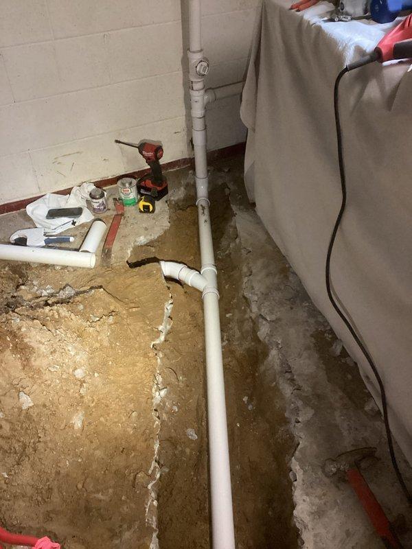 Completed laundry drain installation in basement utility area. Excavated concrete floor trenches to expose substrate, installed white PVC drainage piping with proper vertical and horizontal runs along foundation walls, and routed drainage system to accommodate new laundry equipment location.