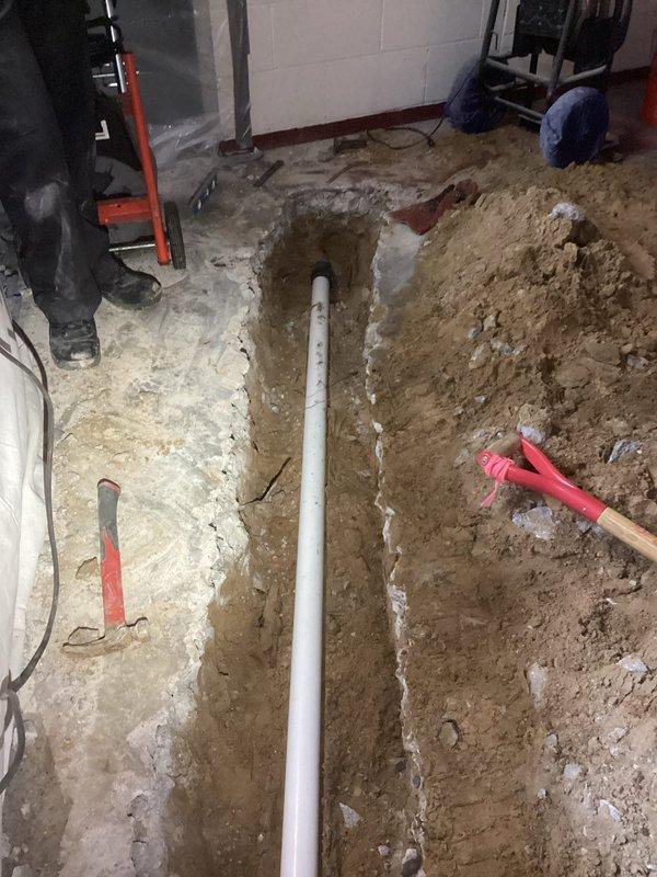 Completed laundry drain installation in basement utility area. Excavated concrete floor trenches to expose substrate, installed white PVC drainage piping with proper vertical and horizontal runs along foundation walls, and routed drainage system to accommodate new laundry equipment location.