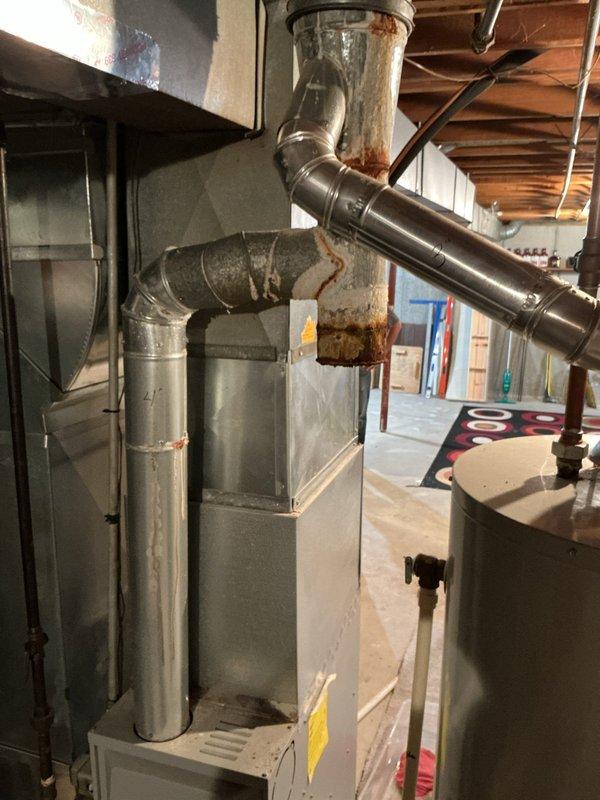 # Heating System Exhaust Flue Inspection

Responded to complaint regarding water intrusion affecting HVAC and water heater exhaust system. Customer reported snow and water entering through secondary chimney flue, causing deterioration of exhaust components.

Upon inspection, confirmed significant rust and corrosion on galvanized metal exhaust ductwork, particularly concentrated at connection points and metal bands where moisture had accumulated. Vertical exhaust pipe showed two distinct bands of severe orange-brown oxidation and deterioration of the galvanized coating. Horizontal branch connection also exhibited weathering and compromised protective coating at junction areas.

Documented current condition of exhaust system with photographs showing extent of corrosion damage on both vertical and horizontal duct sections. Verified exhaust routing through secondary chimney flue. System age could not be determined from available information.

Customer advised of findings regarding water pe