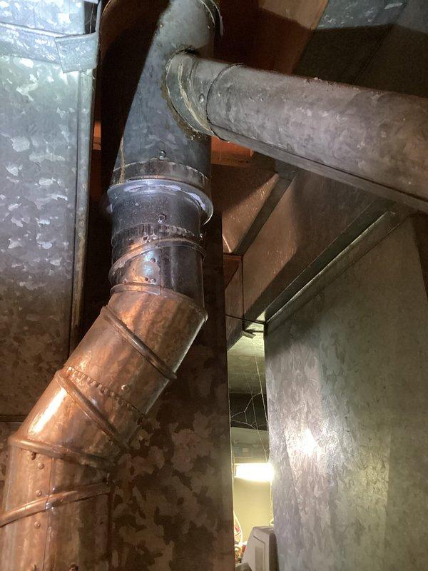 Completed estimate for water heater replacement in residential basement. Assessed existing white cylindrical water heater with connected copper supply and discharge piping, blue valve assembly, and metal exhaust vent system showing signs of age and deterioration. Documented current installation configuration including pipe routing, venting arrangement, and clearance requirements for replacement unit recommendation.