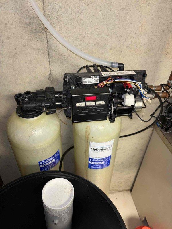 Responded to plumbing leak at residential property with water softener system issue. Upon arrival, found active leak isolated to water softener area in basement utility room. System consists of dual Culligan/Hellenbrand tanks with electronic control valve assembly, previously serviced by our technician. Conducted thorough inspection of 20+ year old equipment and surrounding plumbing connections. Identified source of leak at valve assembly and piping connections. Photographed existing conditions showing discoloration on tank surfaces and control unit configuration. Assessed copper and PVC supply lines, drain connections, and brine tank components. Leak had been present for approximately 24 hours prior to service call. Property covered under home warranty. Documented equipment condition and recommended repair options to resolve active water leak.