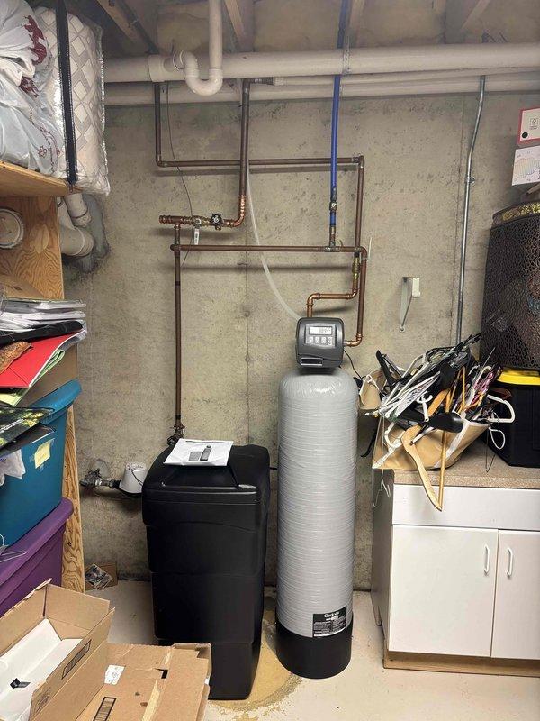 Responded to plumbing leak at residential property with water softener system issue. Upon arrival, found active leak isolated to water softener area in basement utility room. System consists of dual Culligan/Hellenbrand tanks with electronic control valve assembly, previously serviced by our technician. Conducted thorough inspection of 20+ year old equipment and surrounding plumbing connections. Identified source of leak at valve assembly and piping connections. Photographed existing conditions showing discoloration on tank surfaces and control unit configuration. Assessed copper and PVC supply lines, drain connections, and brine tank components. Leak had been present for approximately 24 hours prior to service call. Property covered under home warranty. Documented equipment condition and recommended repair options to resolve active water leak.