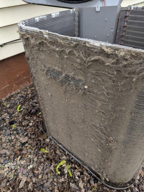 Completed annual cooling maintenance on 9-year-old AC system. Customer reported intermittent galloping sound from basement unit, occurring primarily during peak cooling periods at end of day and occasionally with heating operation, though no consistent pattern identified. Previous technician unable to locate source of noise.

Upon inspection, found outdoor condenser unit with severely restricted airflow due to heavy accumulation of dirt, dust, and organic debris completely caking the aluminum fin coils on multiple sides. Debris buildup was particularly dense on right side and lower portions of coil, significantly reducing heat transfer efficiency and restricting airflow through heat exchanger. This level of contamination would cause reduced cooling capacity, increased operating pressures, and potential compressor strain.

Performed thorough coil cleaning to remove accumulated debris and restore proper airflow through condenser fins. Cleared all obstructions from heat exchange surfaces.
