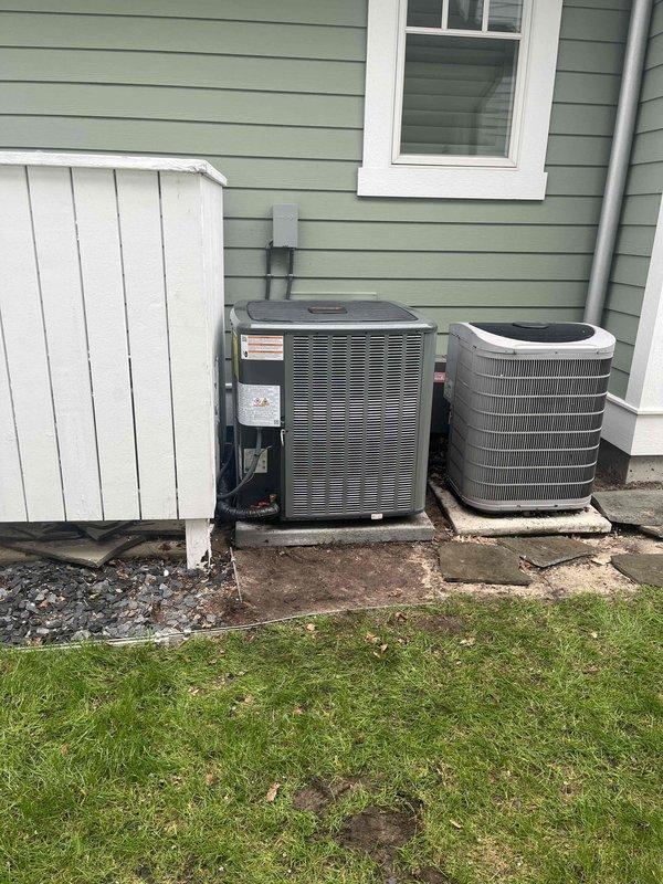 Completed installation of new outdoor HVAC condensing unit at residential property. Unit was properly positioned on concrete pad, with refrigerant lines connected and secured using brass service valves and fittings. Copper line sets were insulated with black ribbed insulation, and all connections were tightened to manufacturer specifications. Electrical conduit was run vertically along exterior wall to provide power supply to the unit. System was charged with refrigerant, pressure tested, and verified operational. Service ports were capped, and unit was labeled with installation data including refrigerant charge information. Second existing unit remains in operation alongside new installation, both positioned on separate concrete pads against home's exterior siding. All work completed per industry standards and manufacturer guidelines.
