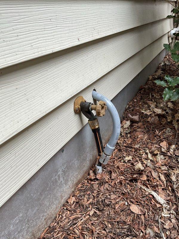 Replaced defective exterior hose bib on building with white lap siding. Removed old brass-colored spigot valve and galvanized curved pipe section that had failed, installed new functioning outdoor faucet assembly with proper threading and secure wall mounting to restore water access to exterior of property.