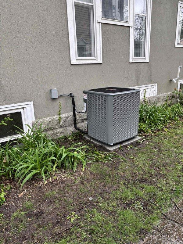 Completed full air conditioning system replacement at residential property. Removed existing outdoor condenser unit and installed new gray HVAC condenser on concrete pad against exterior wall with beige stucco siding. Unit positioned with proper clearance from foundation and windows for adequate airflow and service access. Secured electrical connections via conduit running from wall-mounted disconnect box to condensing unit. Verified unit is level on pad and properly anchored. Tested system operation and confirmed proper refrigerant charge and electrical connections. Work site cleaned and unit operational upon completion.