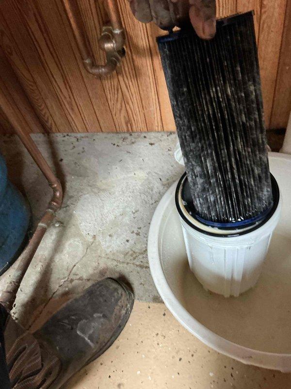 Replaced whole home water filter cartridge due to heavy sediment accumulation and discoloration. Removed old filter element from white cylindrical housing mounted to wooden wall panel, installed new replacement cartridge, and reassembled unit. Verified proper sealing of housing and checked for leaks at copper pipe connections and brass valve fittings. System restored to full operational status with clean filter media in place.