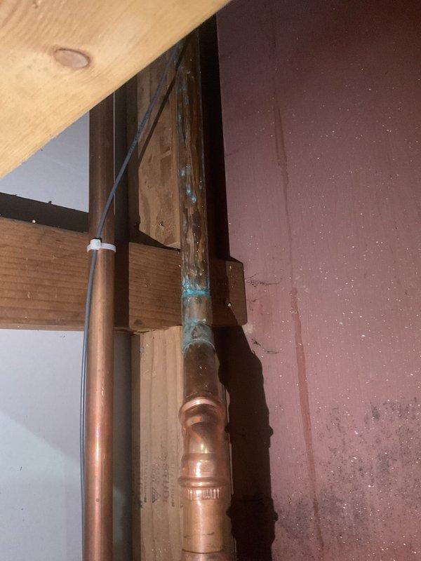 Responded to emergency water leak call originating from copper piping in basement area related to previous installation work. Upon inspection, identified active leak at copper pipe joint with visible blue-green corrosion and discoloration on vertical pipe run within exposed framing structure. Assessed compromised solder joint connection and copper elbow fitting in crawl space area. Repaired defective pipe connection, addressed corrosion issue, and restored system integrity. Verified repair under pressure and confirmed no additional leaks present. Documented conditions with photos for service records.
