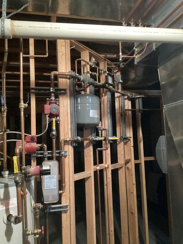 Completed annual cooling system maintenance and inspection. Documented mechanical room installation showing hydronic heating components, circulation pumps, expansion tanks, and copper piping distribution system in unfinished space with exposed framing. All photographed equipment and connections verified for proper operation and accessibility.
