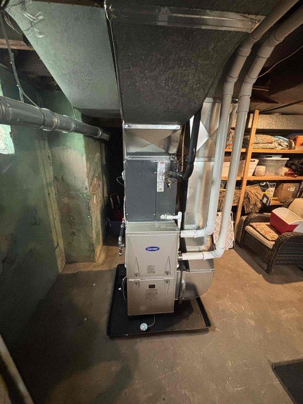 Completed full furnace installation in basement mechanical room, installing new Carrier brand furnace unit on protective platform with proper ventilation system including white PVC exhaust and intake piping routed through ceiling structure. Connected gray metal ductwork to existing home ventilation system and verified all connections secure and operational.