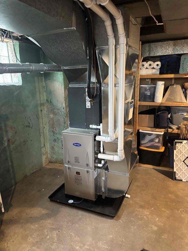 Completed full furnace installation in basement mechanical room, installing new Carrier brand furnace unit on protective platform with proper ventilation system including white PVC exhaust and intake piping routed through ceiling structure. Connected gray metal ductwork to existing home ventilation system and verified all connections secure and operational.