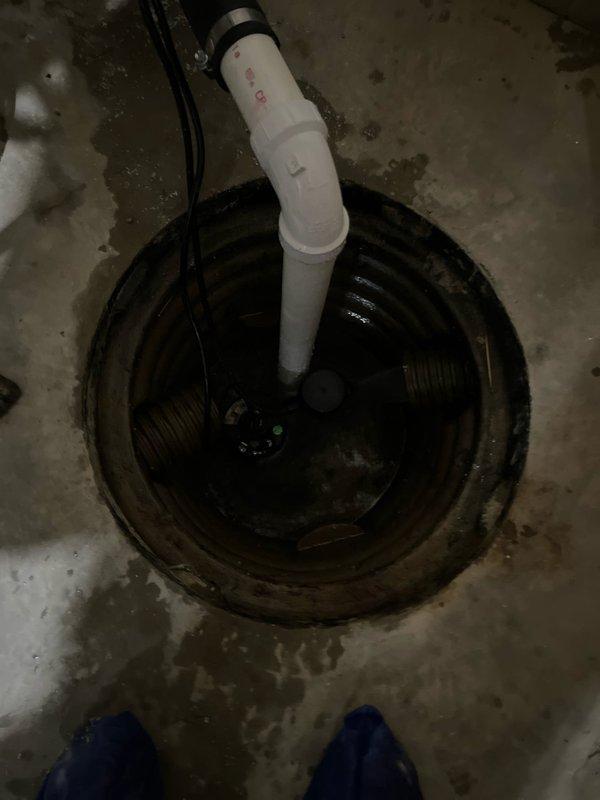 Responded to service call for non-operational sump pump in 20-year-old residential basement. Upon inspection, found sump pump system with vertical PVC discharge pipe and standing water in corrugated plastic pit, along with evidence of moisture staining on surrounding concrete floor indicating drainage concerns. Documented existing installation configuration including pump assembly, electrical connections, and discharge piping for assessment and resolution of operational failure.
