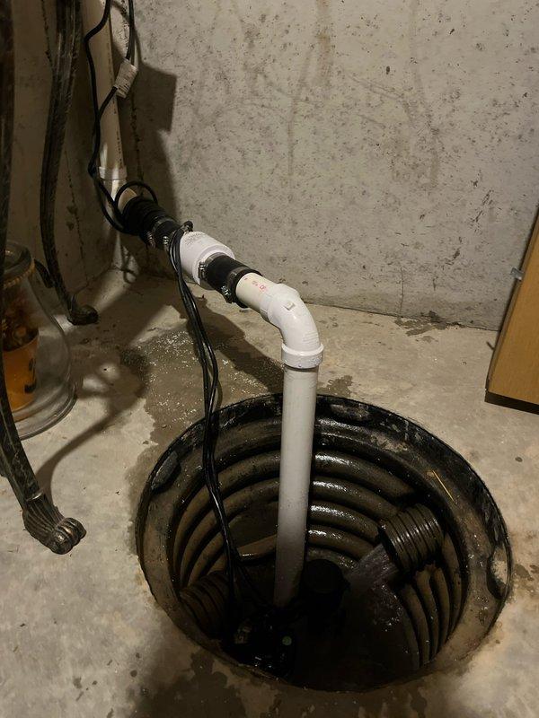 Responded to service call for non-operational sump pump in 20-year-old residential basement. Upon inspection, found sump pump system with vertical PVC discharge pipe and standing water in corrugated plastic pit, along with evidence of moisture staining on surrounding concrete floor indicating drainage concerns. Documented existing installation configuration including pump assembly, electrical connections, and discharge piping for assessment and resolution of operational failure.