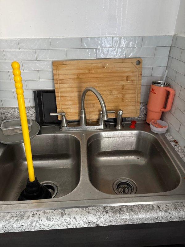 Responded to clogged kitchen sink in residential property. Upon arrival, found standing water in left basin of stainless steel double-basin sink. Used plunger to attempt initial clearance of drain blockage. Successfully cleared obstruction from kitchen drain line, restoring proper water flow and drainage to both sink basins. Tested system to confirm complete resolution of clog and verified no leaks present.