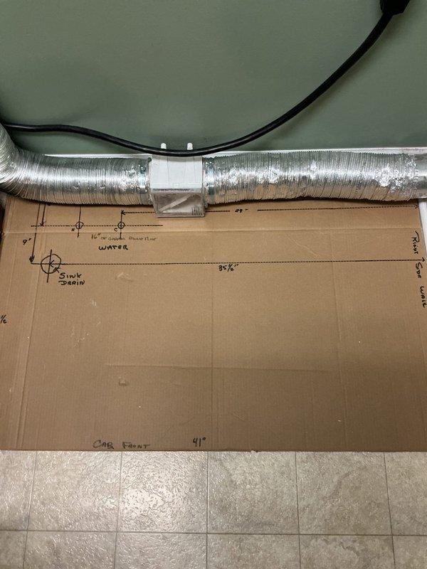 Conducted on-site plumbing estimate for laundry room sink installation. Assessed existing space and documented precise measurements for water supply and drain line routing through floor. Created detailed dimensional template on cardboard indicating sink placement, cabinet front positioning, water line location, and drain pipe penetration point. Evaluated proximity to existing HVAC ductwork and electrical components to determine optimal routing path. Photographed workspace configuration and measurement layout for accurate material procurement and installation planning.