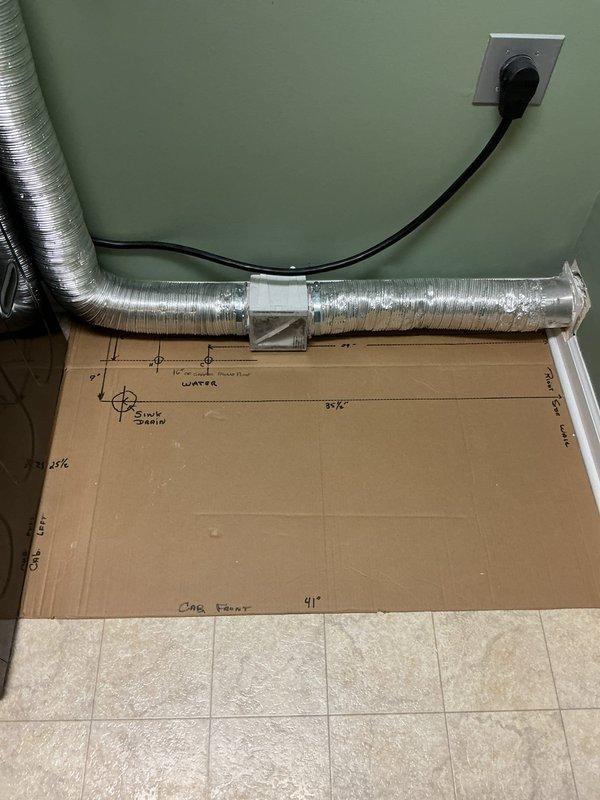 Conducted on-site plumbing estimate for laundry room sink installation. Assessed existing space and documented precise measurements for water supply and drain line routing through floor. Created detailed dimensional template on cardboard indicating sink placement, cabinet front positioning, water line location, and drain pipe penetration point. Evaluated proximity to existing HVAC ductwork and electrical components to determine optimal routing path. Photographed workspace configuration and measurement layout for accurate material procurement and installation planning.