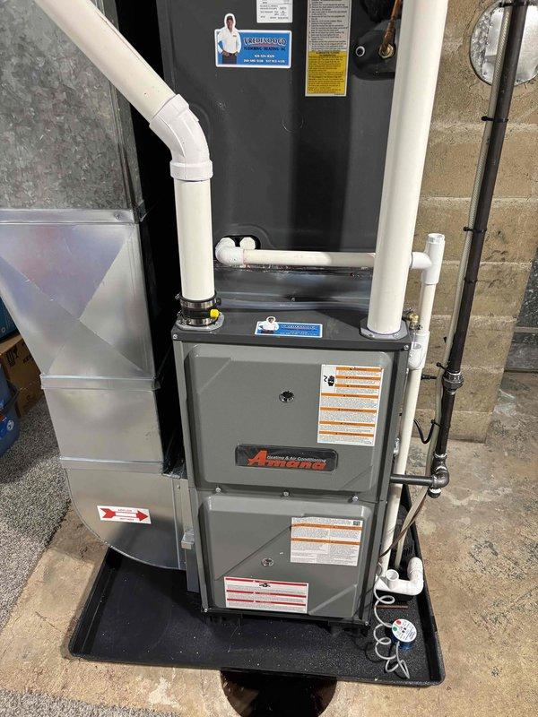 Completed full furnace installation in basement utility area. Installed gray Amana furnace unit on black drain pan with proper positioning on concrete floor against concrete block wall. Connected white PVC ventilation and condensate drainage piping from furnace top, integrated system with existing air handler and ductwork components. Verified all manufacturer labels, safety specifications, and operating instructions are visible and properly affixed to unit.