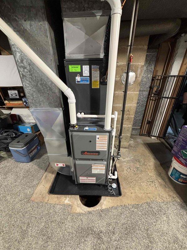 Completed full furnace installation in basement utility area. Installed gray Amana furnace unit on black drain pan with proper positioning on concrete floor against concrete block wall. Connected white PVC ventilation and condensate drainage piping from furnace top, integrated system with existing air handler and ductwork components. Verified all manufacturer labels, safety specifications, and operating instructions are visible and properly affixed to unit.