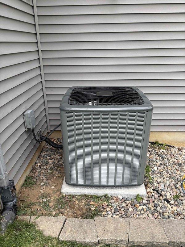 Completed full air conditioning system replacement at residential property. Removed existing outdoor condensing unit and installed new AC unit on concrete pad against exterior wall with proper electrical disconnect and conduit installation. Unit positioned on white concrete pad with surrounding river rock landscaping, ensuring proper clearances and drainage for optimal system performance.