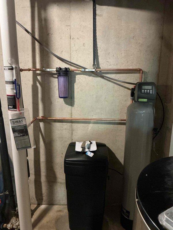 Completed full water softener system installation in basement utility area. Installed black water softener tank with digital control head and gray brine tank with digital display panel, connected via copper piping network with bypass configuration. Mounted purple water filter housing on horizontal pipe run above tanks, integrated all components with existing plumbing system including water heater connection. Installation took approximately 4 hours to complete with all equipment properly positioned, piped, and tested for operation.