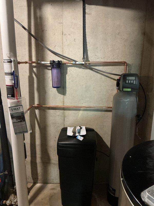 Completed full water softener system installation in basement utility area. Installed black water softener tank with digital control head and gray brine tank with digital display panel, connected via copper piping network with bypass configuration. Mounted purple water filter housing on horizontal pipe run above tanks, integrated all components with existing plumbing system including water heater connection. Installation took approximately 4 hours to complete with all equipment properly positioned, piped, and tested for operation.