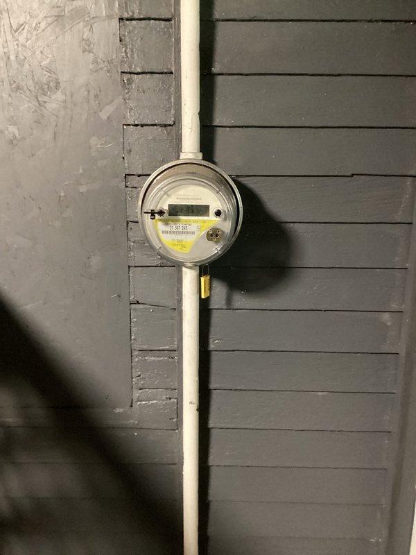 Conducted electrical assessment and provided estimate for 96-year-old residential property following recent inspection findings. Evaluated existing exterior electrical meter assembly mounted on dark gray horizontal siding with white conduit system and visible inspection tag. Assessed interior electrical infrastructure including exposed basement/crawl space areas with aging electrical components and PVC conduit routing through wooden joists. Documented current electrical panel condition, meter configuration, and overall system integrity to determine scope of work required for electrical box replacement and additional necessary upgrades to bring installation into compliance with current code requirements.