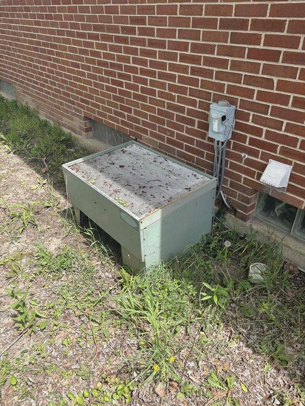 Responded to service call for HVAC system evaluation and relocation estimate. Upon arrival, inspected outdoor condensing unit positioned alongside building exterior. Unit displayed significant accumulation of dirt, dust, and debris on condenser coils and casing, with heavy layer of grime obscuring metal surfaces. Condenser exhibited cylindrical design with spiral coil pattern typical of older equipment configuration. System appeared to be over 10 years in service based on visible wear and weathering patterns.

Conducted thorough visual assessment of unit placement and surrounding area. Equipment currently situated on ground-level pad adjacent to red brick exterior wall with electrical disconnect box and conduit properly mounted. Noted vegetation growth around unit base and debris accumulation on top surface indicating extended exposure to outdoor elements without recent maintenance.

Documented current installation location and condition through photographic evidence for relocation est