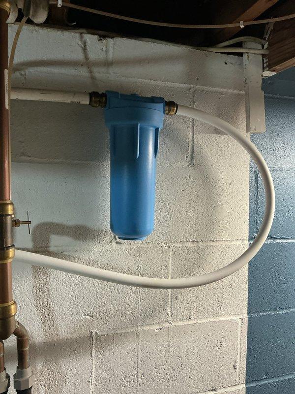 Responded to emergency water leak call and located source at whole-house water filtration system in basement utility area. Upon inspection, identified leak originating from compromised connection points at brass fittings on blue cylindrical filter housing mounted to concrete block wall. White plastic supply tubing showed signs of stress at compression fittings where water was actively seeping. 

Shut off main water supply to prevent further water damage and assessed the filtration system installation. Disassembled the affected brass compression fittings at both inlet and outlet connections on the filter housing. Inspected tubing ends for damage and cut back sections showing stress marks to ensure clean, square cuts for proper seal. Replaced worn ferrules and compression rings that had lost their sealing capability.

Reassembled all connections with new compression components, ensuring proper torque specifications to prevent over-tightening while achieving watertight seal. Restored wate
