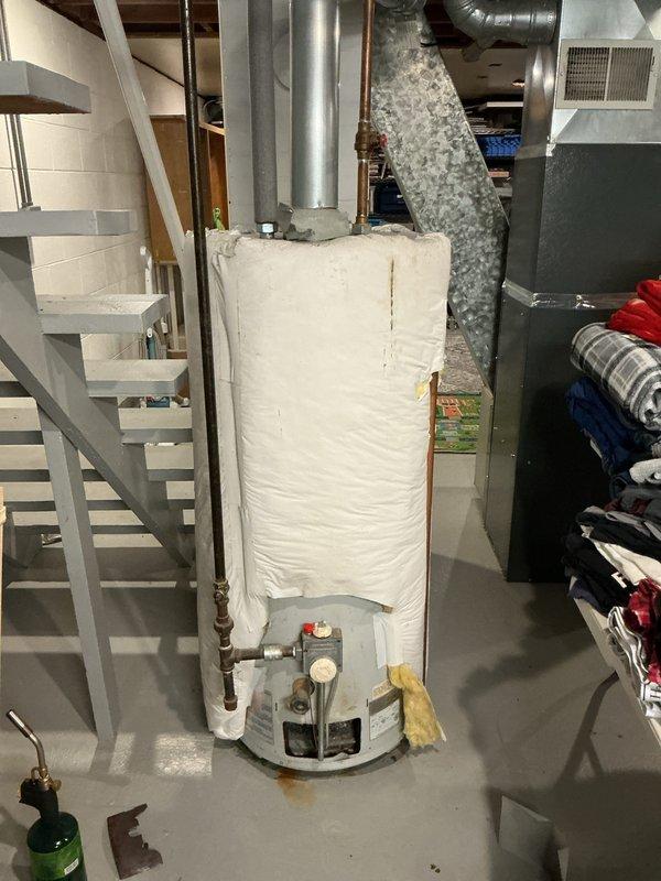 # Water Heater Service Call

Responded to service call for non-operational water heater approximately 10 years old. Upon arrival, conducted visual inspection of gas water heater unit located in basement utility area. Examined unit wrapped in white insulation blanket, assessed plumbing connections including copper supply lines and shut-off valves, and inspected venting system with metallic duct extending from top of unit. Noted rust-colored staining on concrete floor around base of water heater, indicating potential previous leak or condensation issues. Checked temperature and pressure relief valve, pressure gauge, and control mechanisms at base of unit. Documented existing conditions with photographs for service records. Unit positioned in close proximity to HVAC furnace system with shared utility space.