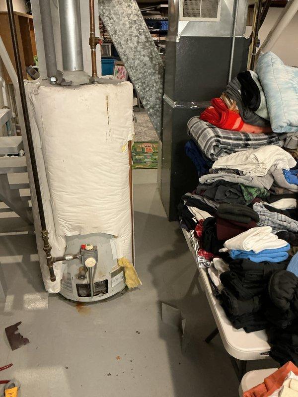 # Water Heater Service Call

Responded to service call for non-operational water heater approximately 10 years old. Upon arrival, conducted visual inspection of gas water heater unit located in basement utility area. Examined unit wrapped in white insulation blanket, assessed plumbing connections including copper supply lines and shut-off valves, and inspected venting system with metallic duct extending from top of unit. Noted rust-colored staining on concrete floor around base of water heater, indicating potential previous leak or condensation issues. Checked temperature and pressure relief valve, pressure gauge, and control mechanisms at base of unit. Documented existing conditions with photographs for service records. Unit positioned in close proximity to HVAC furnace system with shared utility space.