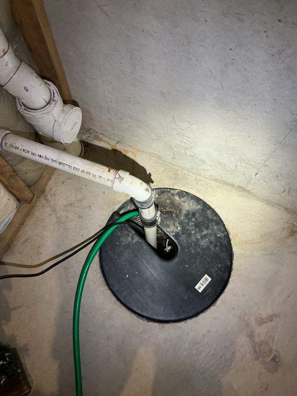 Completed comprehensive Plumbing Club Inspection documenting existing plumbing conditions. Identified and photographed damaged white PVC pipe with visible break and rough torn edges in utility area, along with metal plumbing fixture assembly featuring braided flexible hoses. Documented sump pump installation including basin cover, discharge piping, and associated PVC plumbing components in basement area for maintenance records.