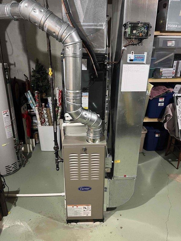 Complete HVAC system installation performed at residential property. Installed new Carrier furnace unit in basement utility area with corresponding ductwork configuration, including galvanized metal supply ducts with proper support strapping and elbow transitions. Exterior condensing unit positioned on concrete pad against house exterior with tan vinyl siding, connected via refrigerant lines and electrical conduit routed to interior equipment. Air handler cabinet installed adjacent to furnace with electrical junction box mounted and properly wired. All equipment secured, leveled, and tested for proper operation. System commissioned and verified operational parameters within manufacturer specifications.