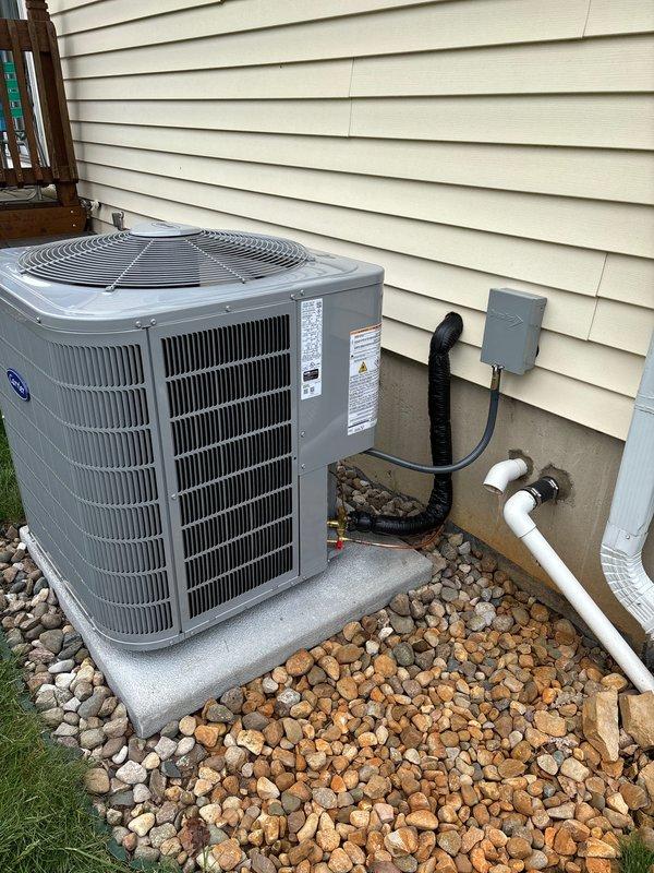 Completed full air conditioning system replacement including outdoor condensing unit and indoor air handler. Installed new gray cylindrical condenser on existing concrete pad with proper clearances for airflow, connected electrical disconnect box with conduit, and routed white PVC condensate drain lines along exterior wall. Replaced indoor air handler and metal ductwork plenum assembly in basement utility area. Verified all electrical connections, refrigerant lines, and condensate drainage systems functioning properly. System tested and operational upon completion.