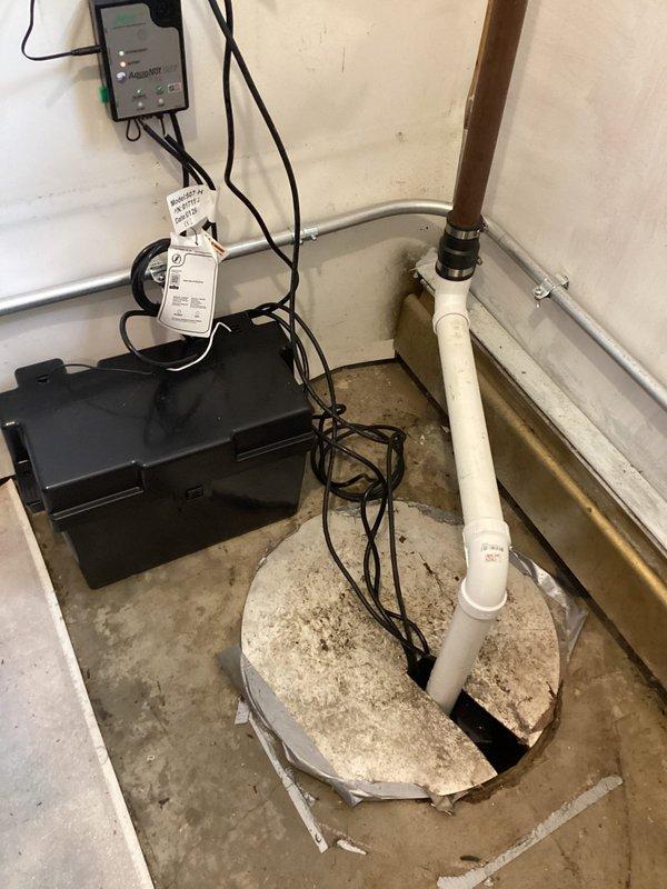 Completed installation of battery backup sump pump system in basement location. Installed primary sump pump in concrete pit with PVC discharge piping running vertically to overhead connection point. Mounted wall-mounted control panel with indicator lights and installed floor-level battery backup unit with all necessary electrical connections and cabling between components. System tested and operational, providing automatic backup protection during power outages.