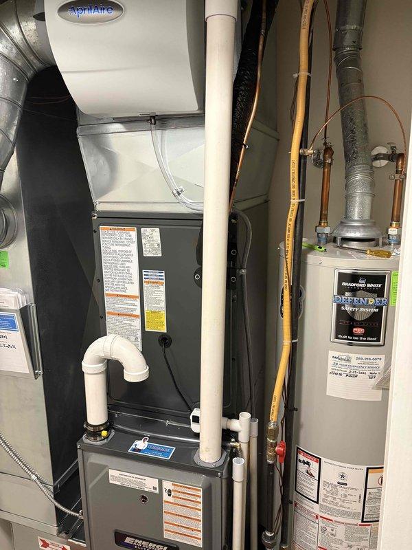 Completed full installation of new residential HVAC system including furnace and air conditioning condenser unit. Furnace installed in mechanical room with proper venting via white PVC piping, integrated with AprilAire humidifier system, and connected to existing ductwork. Outdoor AC condenser positioned on concrete pad in side yard with appropriate clearance from structure, refrigerant lines installed with black foam insulation running through exterior wall connection. All equipment properly labeled with manufacturer specifications and safety warnings, system tested and operational.