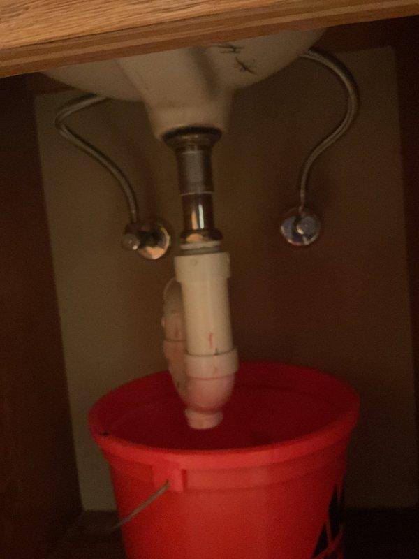Responded to recurring plumbing leak beneath kitchen sink after multiple previous service calls for the same issue. Upon arrival, inspected the PVC P-trap assembly and drain connections where active leaking had been reported. Placed collection bucket under the drain system to catch water and prevent cabinet damage while assessing the problem. Examined all pipe connections, tailpiece fitting, and P-trap joints for signs of deterioration, improper sealing, or structural failure. Documented the plumbing configuration with photographs showing the white PVC P-trap, metal tailpiece connection, flexible supply lines, and cabinet interior condition. No trip charge was applied per prior authorization due to the repeat nature of this service call.