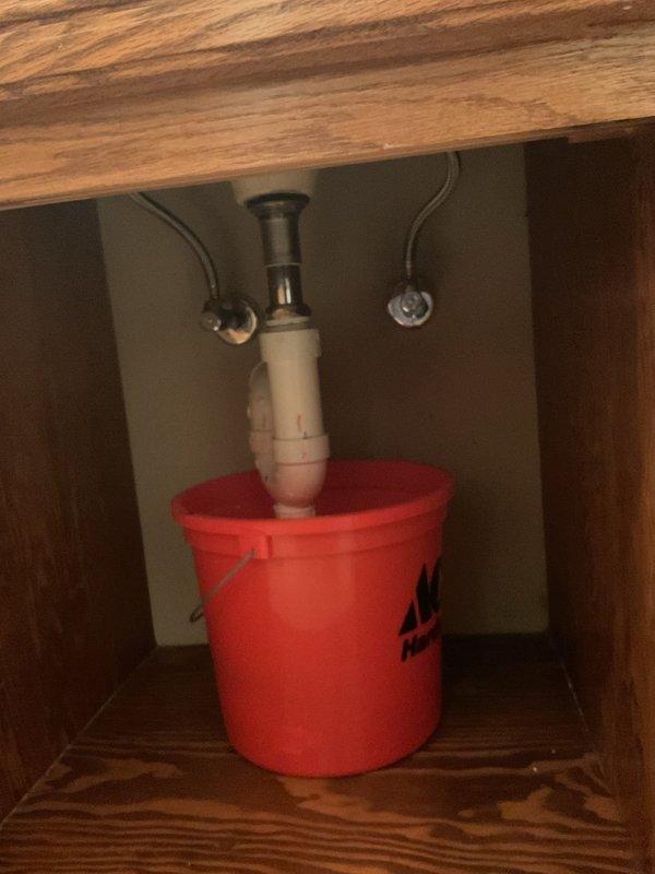 Responded to recurring plumbing leak beneath kitchen sink after multiple previous service calls for the same issue. Upon arrival, inspected the PVC P-trap assembly and drain connections where active leaking had been reported. Placed collection bucket under the drain system to catch water and prevent cabinet damage while assessing the problem. Examined all pipe connections, tailpiece fitting, and P-trap joints for signs of deterioration, improper sealing, or structural failure. Documented the plumbing configuration with photographs showing the white PVC P-trap, metal tailpiece connection, flexible supply lines, and cabinet interior condition. No trip charge was applied per prior authorization due to the repeat nature of this service call.